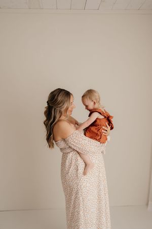 A woman in a floral off-shoulder dress holds a small child wearing an orange outfit against a neutral background.