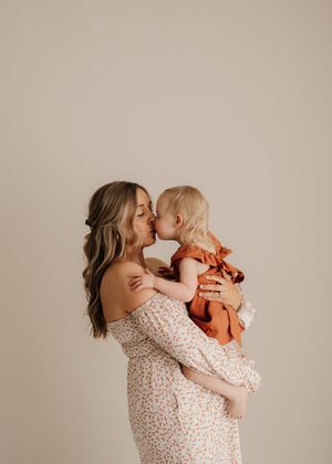 Tender moment showing woman in floral off-shoulder dress holding and kissing young child in orange outfit against beige wall.