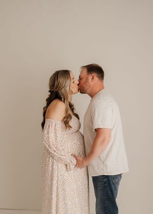 A couple in casual white outfits express affection through an intimate kiss against a neutral studio backdrop.