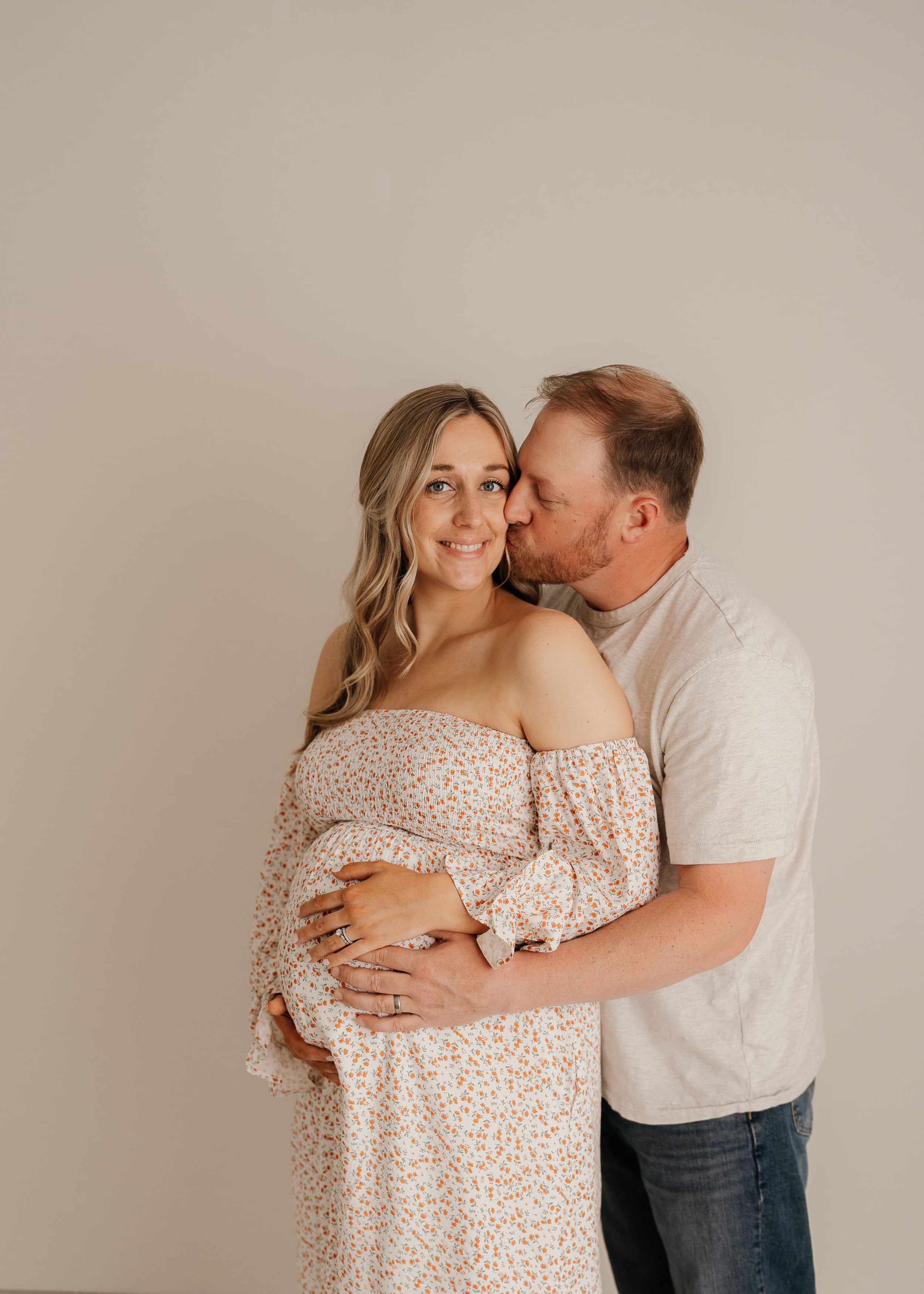 Couple shares an intimate moment during a maternity photoshoot against a neutral beige background.