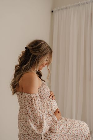 A pregnant woman in a floral off-shoulder dress poses near a window with soft natural light.