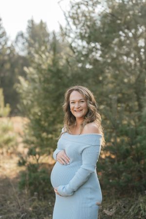 Expecting mother in light blue dress smiles while standing in natural outdoor setting.