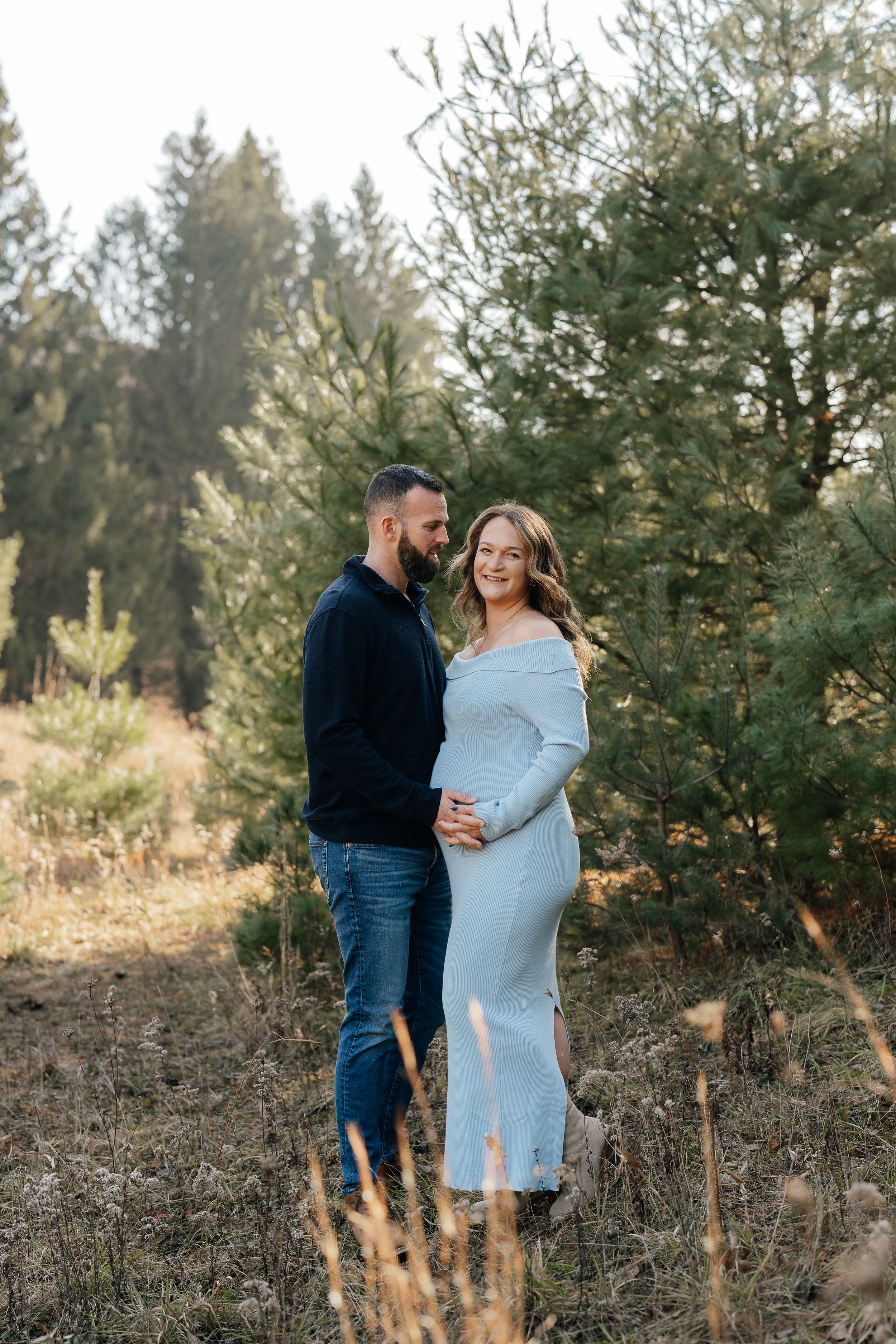 Expectant couple embraces in a rustic outdoor setting among evergreen trees.