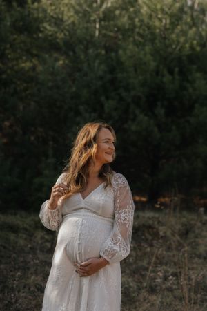 Expectant mother in white lace dress poses against forest backdrop during golden hour.