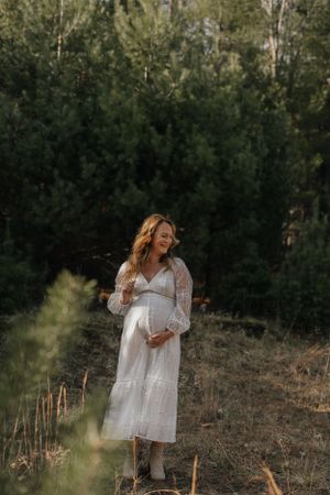 Maternity photo of woman in white dress standing on hillside with pine forest background.