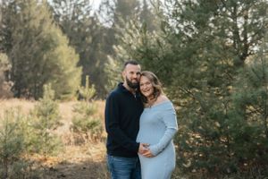 Intimate maternity photo of couple standing among pine trees at sunset.