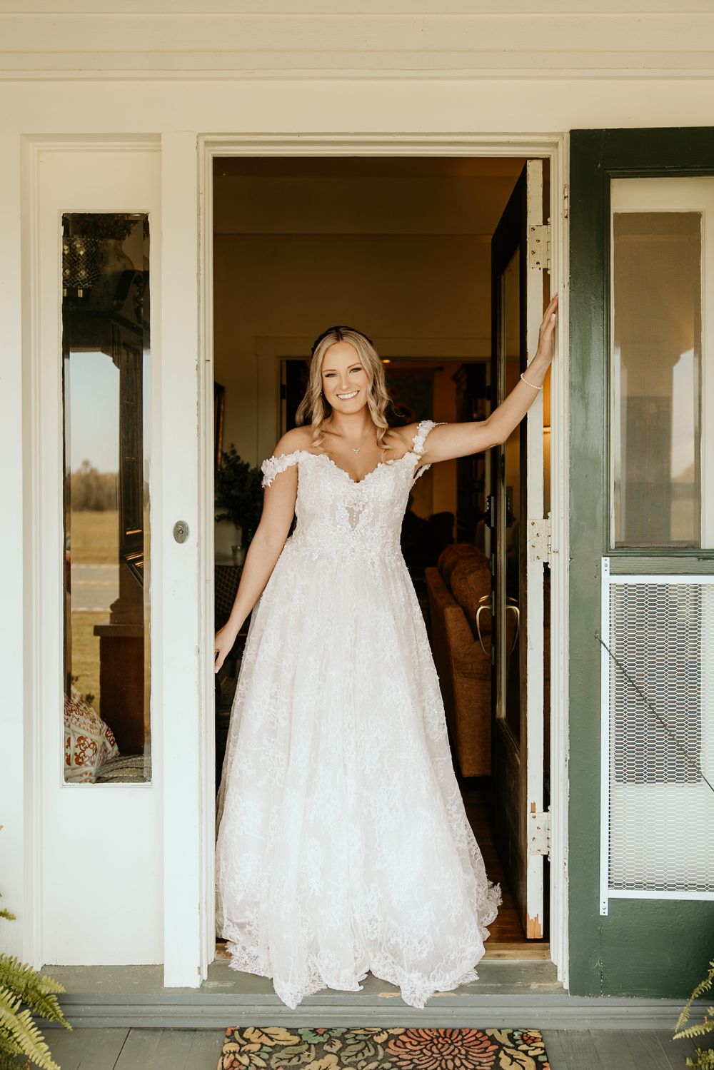 Photograph of Bride in elegant lace wedding gown stands in doorway of white farmhouse at Wild Daisy Farm in Molena, Georgia.
