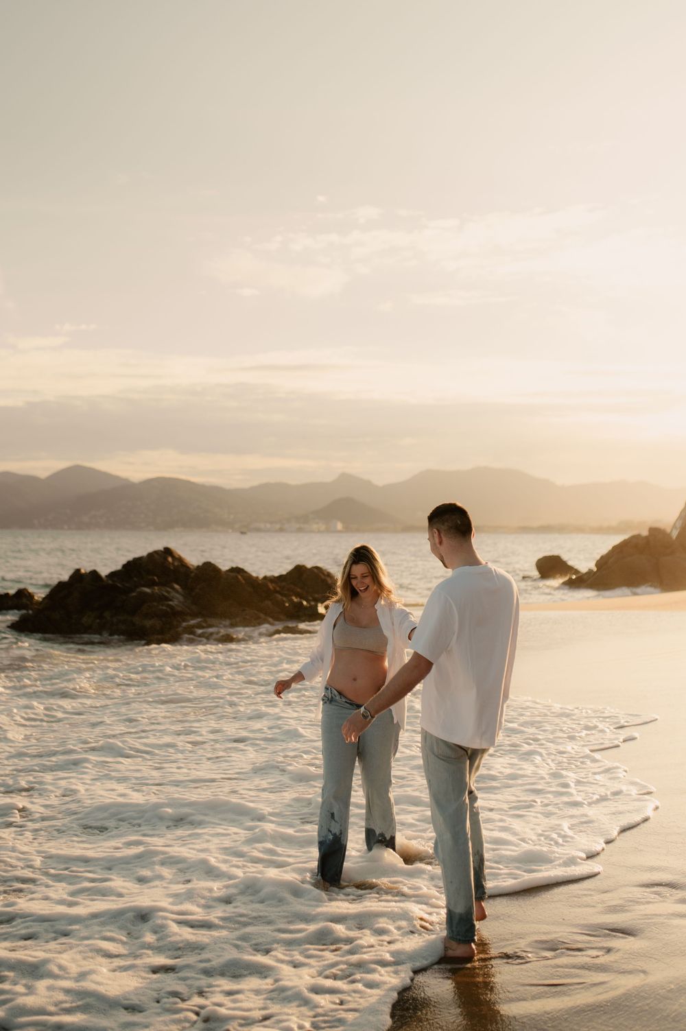 Séance photo d'un jeune couple qui attendent un bébé - Ils marchent sous une lumière pastel, dans la mer et les vagues