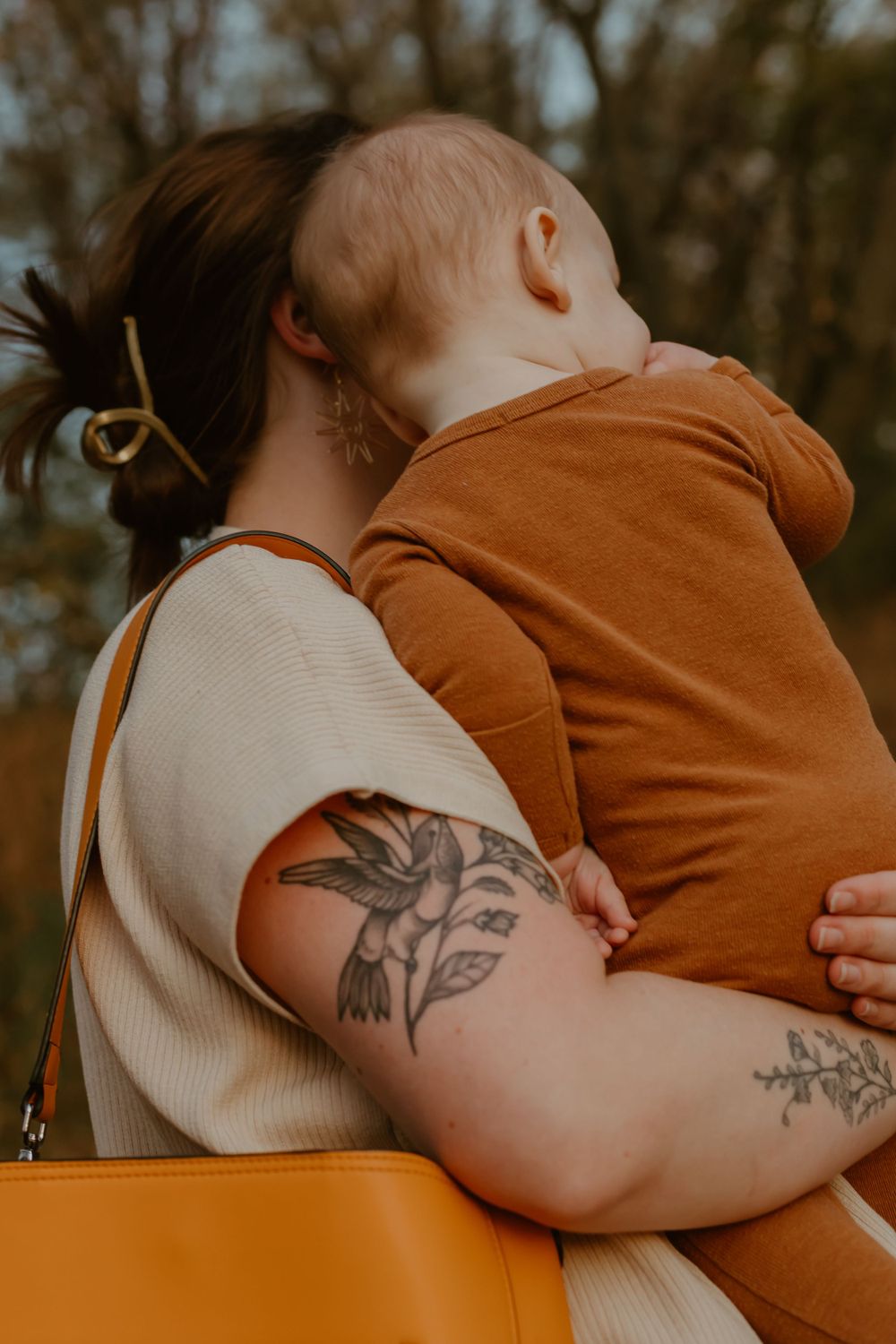 Close-up view of a tattoo-adorned arm holding a child wearing orange against golden autumn trees.