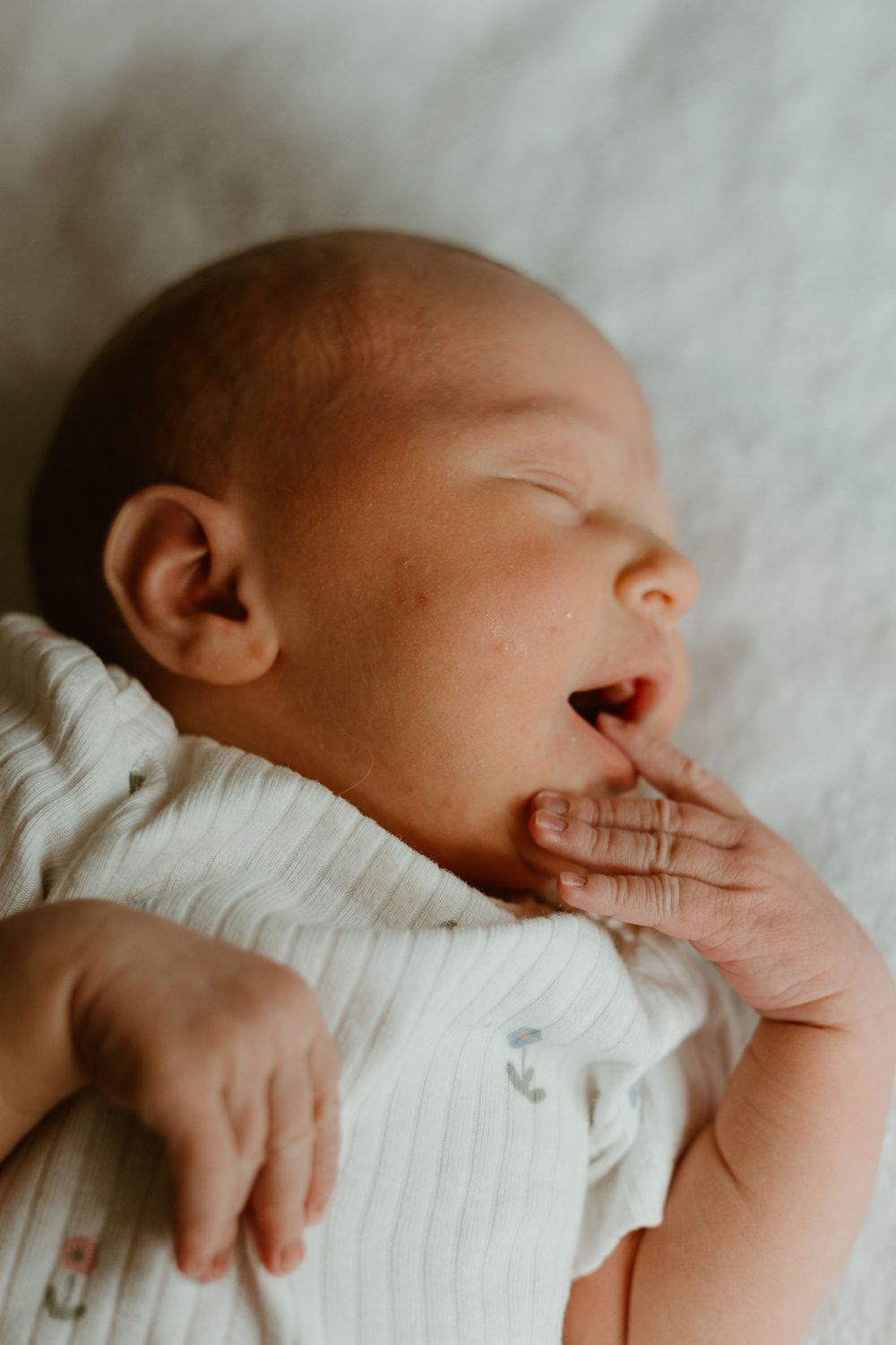 A sleepy newborn baby wearing a white cardigan stretches and yawns while resting on white bedding.