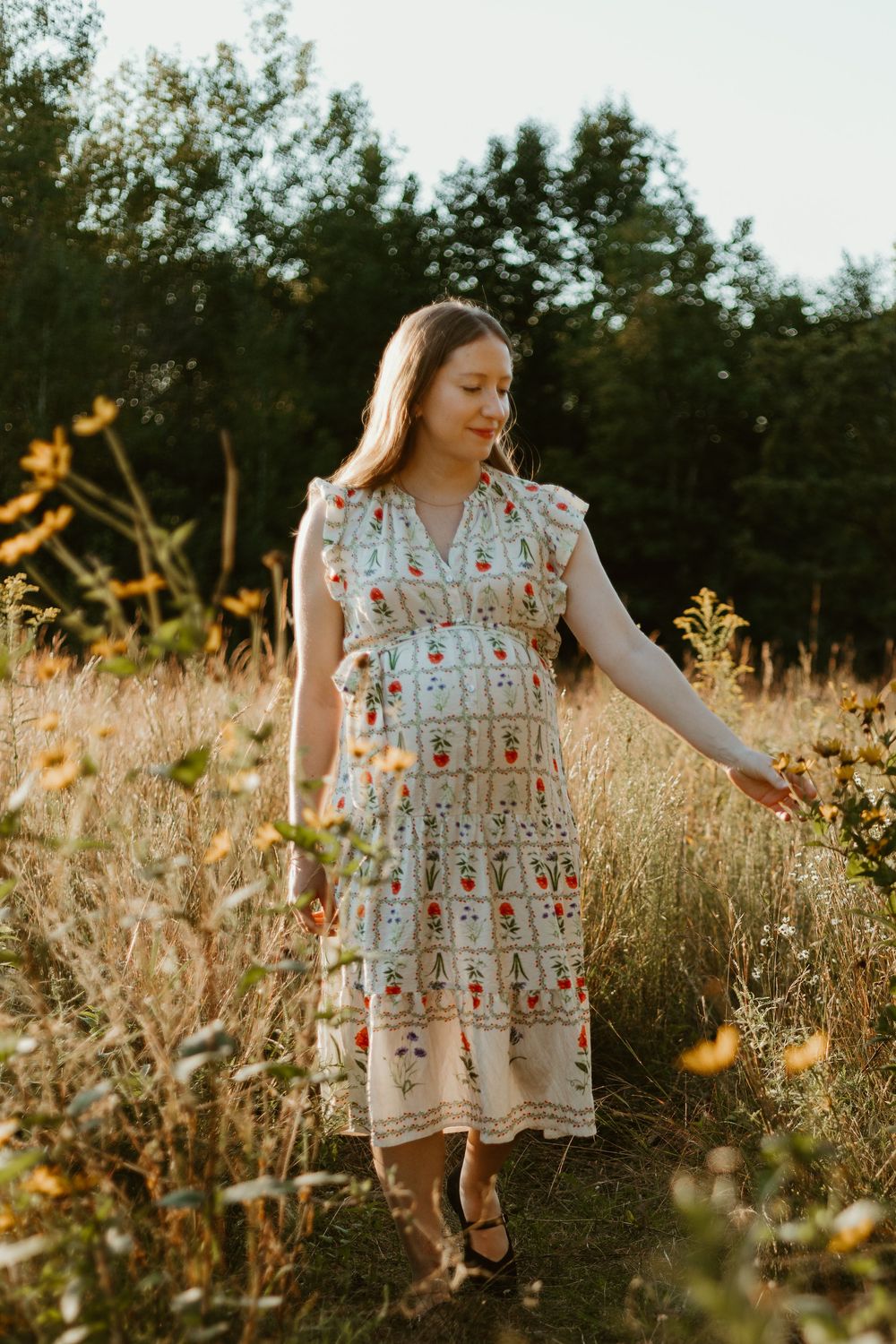 A woman in a floral embroidered dress walks through a golden meadow at sunset with wildflowers in bloom.