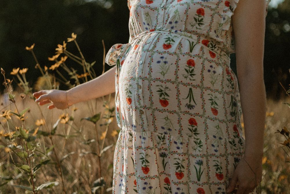 Woman in a floral white summer dress poses in a golden field during sunset, creating a dreamy ethereal scene.
