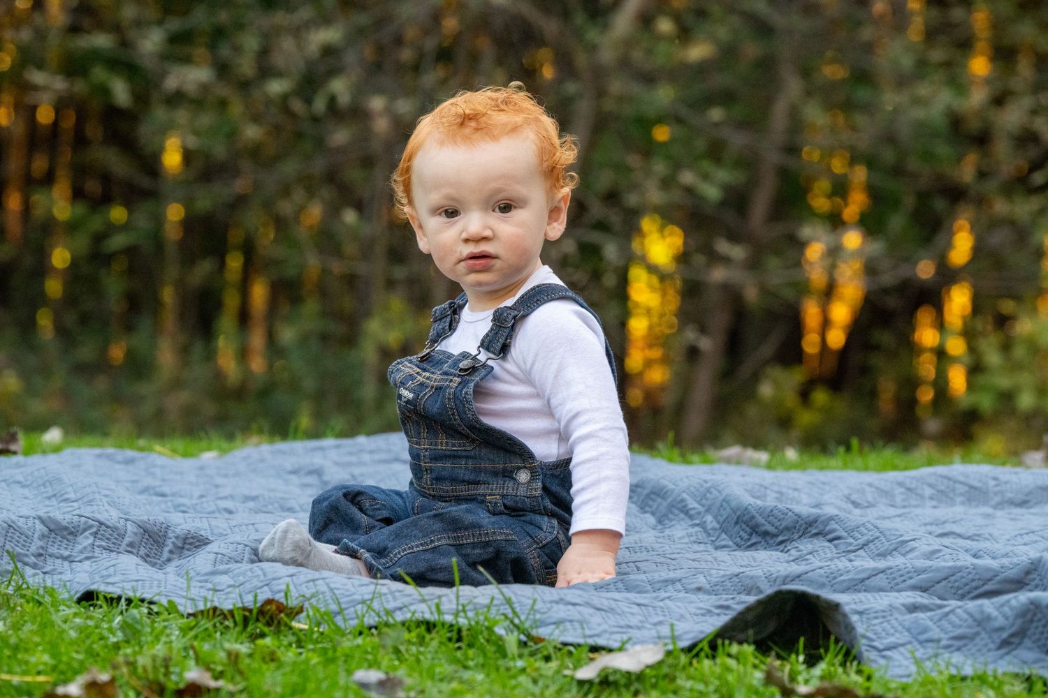 A baby sits on a blue blanket outdoors surrounded by autumn foliage.