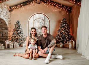 Family sitting on floor in front of decorated Christmas trees and twinkling lights in cozy holiday setting.