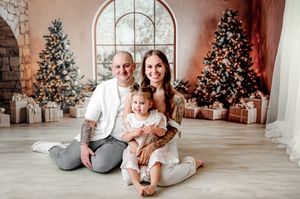 Family of three posing together in front of decorated Christmas trees with gifts in a festive holiday photo session.