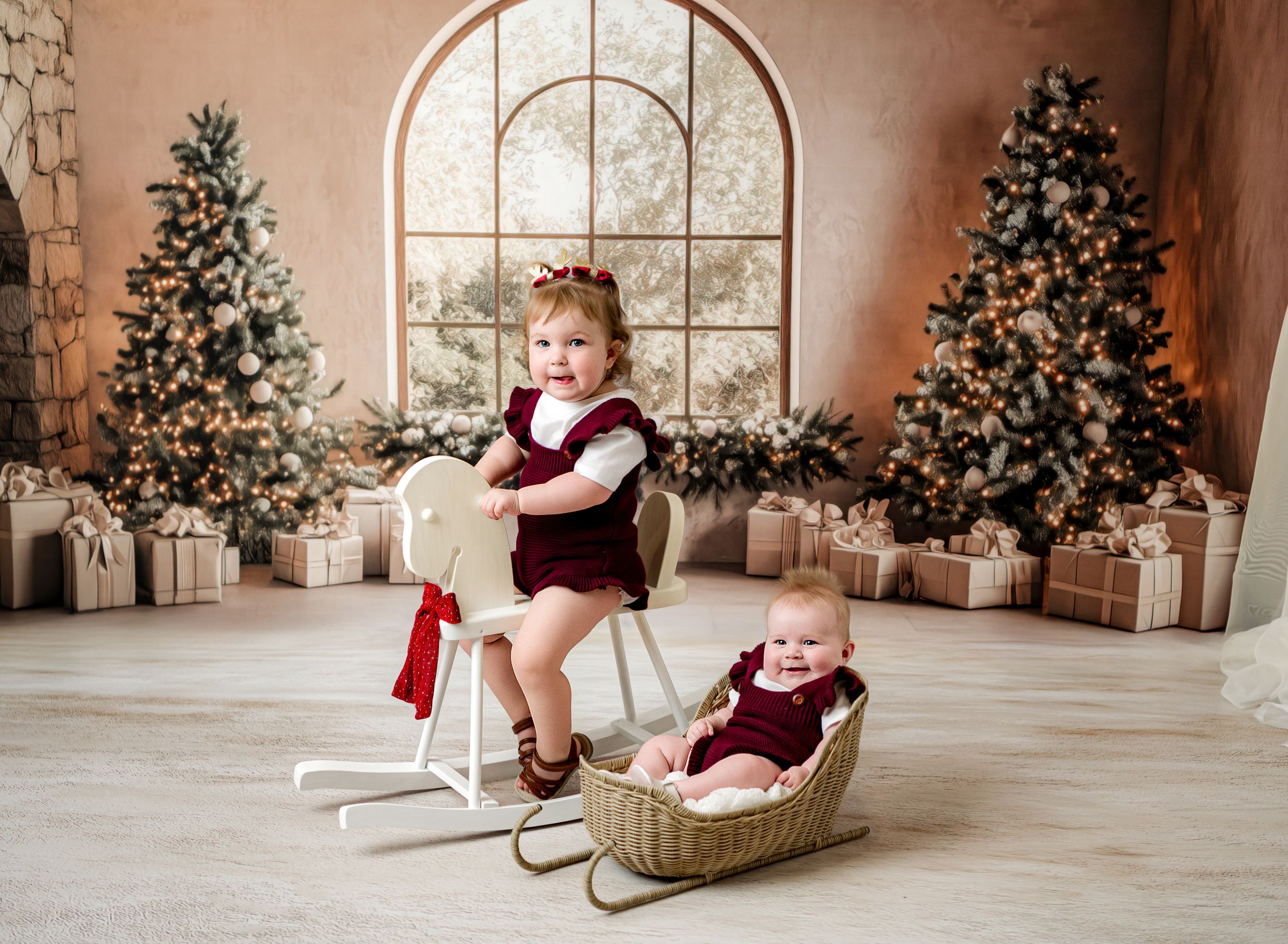 Two young children in burgundy outfits pose with Christmas trees and gifts in a festive holiday photo studio setting.