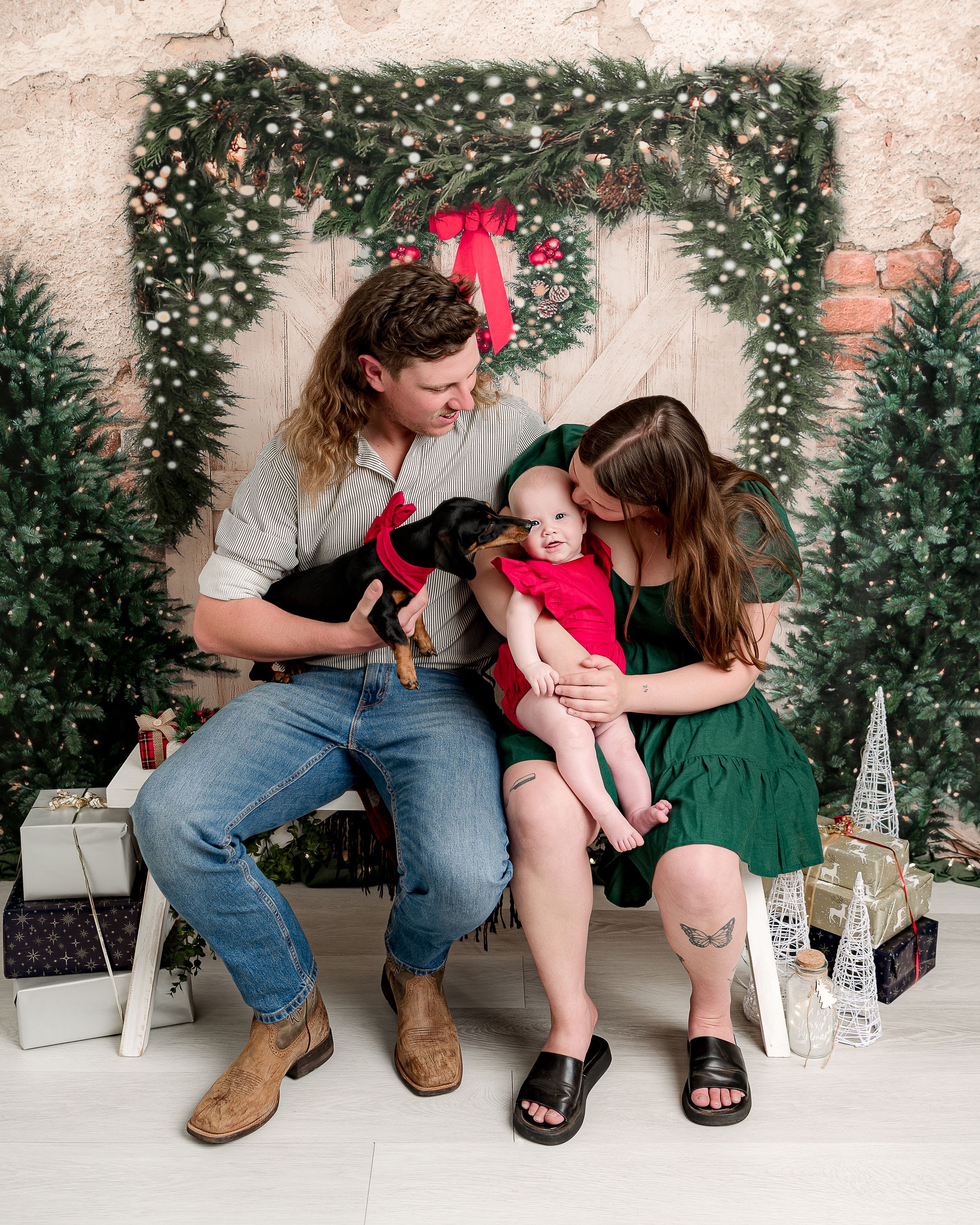 Loving family moment captured against holiday backdrop with greenery and red bow wreath.