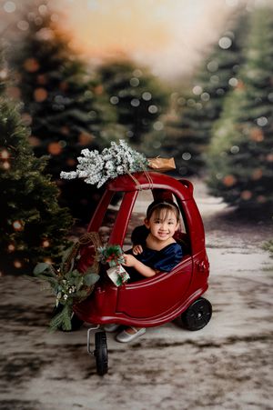 A smiling child sits in a red toy car with a Christmas tree on top, surrounded by a festive tree farm backdrop.