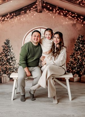 Family of three poses for Christmas portrait with decorated trees and warm string lights in cozy studio.