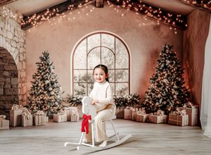 Young girl on white rocking horse in festive Christmas studio with decorated trees and wrapped gifts.