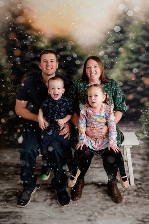 Family of four poses for a festive Christmas photo with snowy backdrop and holiday trees.