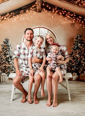 Family of four in matching plaid Christmas pajamas posing on bench with decorated trees and festive lights backdrop.