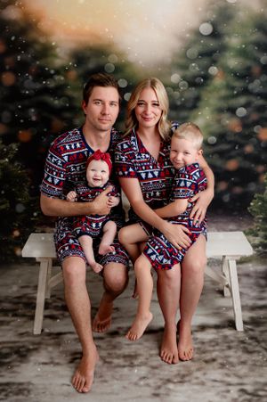 Family wearing matching Christmas pajamas poses together on white bench amid snowy scene.