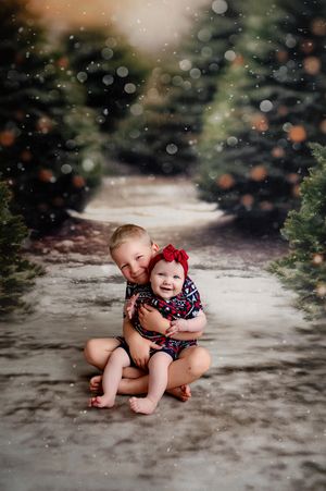 Two young siblings hugging among Christmas trees with falling snow in a festive holiday portrait photo.