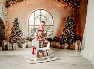 Cozy Christmas photo studio with decorated trees, arched windows, rocking horse, and warm glowing lights.