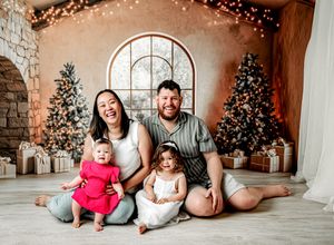Family of four poses joyfully before decorated Christmas trees with twinkling lights and wrapped gifts in a festive studio setting.