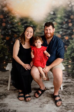 Family of three posing for a festive Christmas portrait with bokeh lights and snow-dusted pine trees in the background.