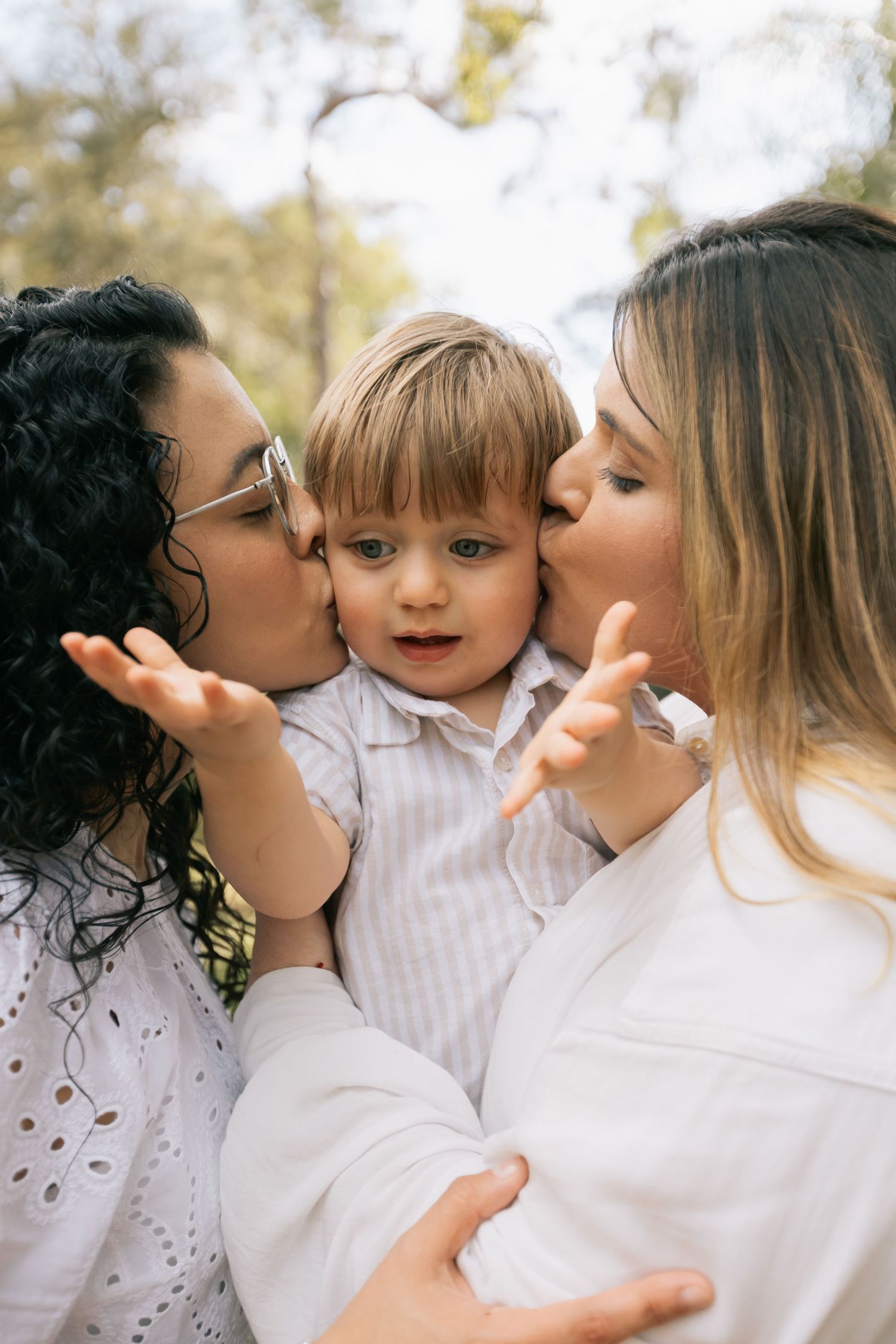 Two women kiss the cheeks of a smiling toddler boy in a striped shirt while he reaches out his small hands outdoors.