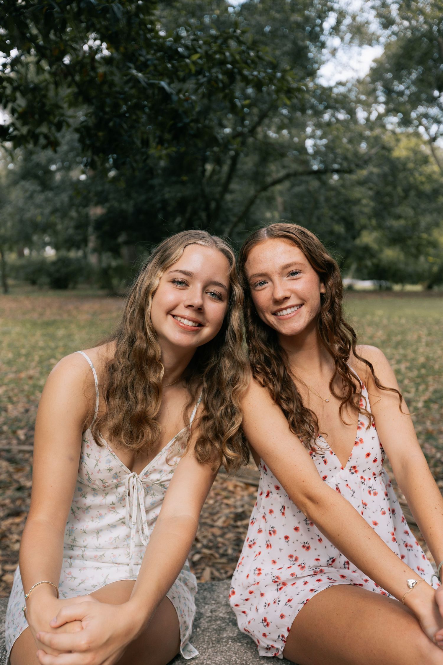 Two smiling young women with long wavy hair sit close together in floral sundresses on a grassy park lawn beneath tall trees.