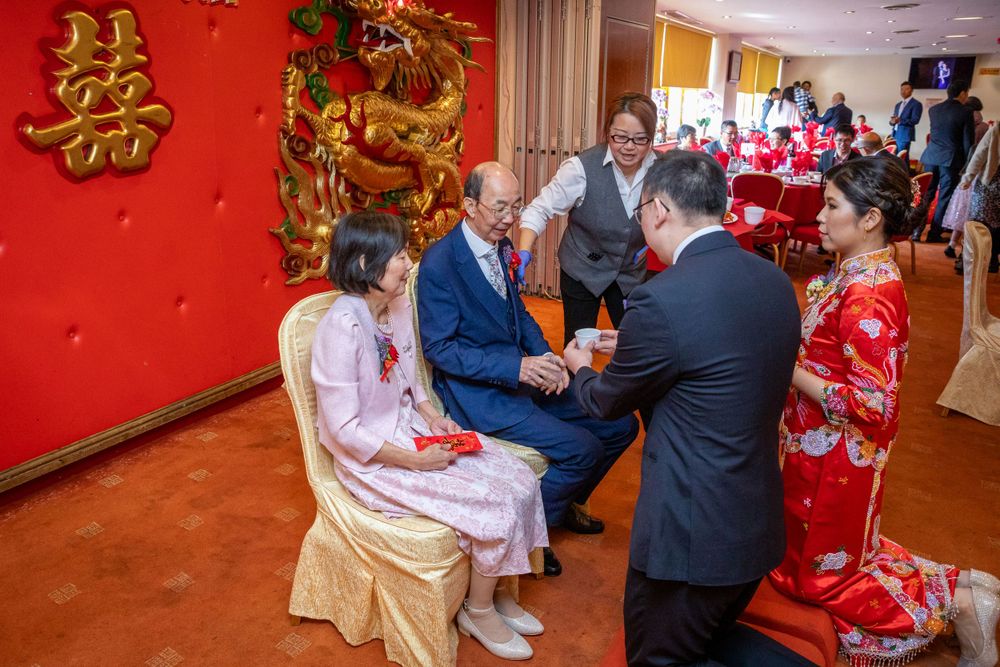Couple performing traditional Chinese wedding tea ceremony serving elders at a red-decorated banquet venue.