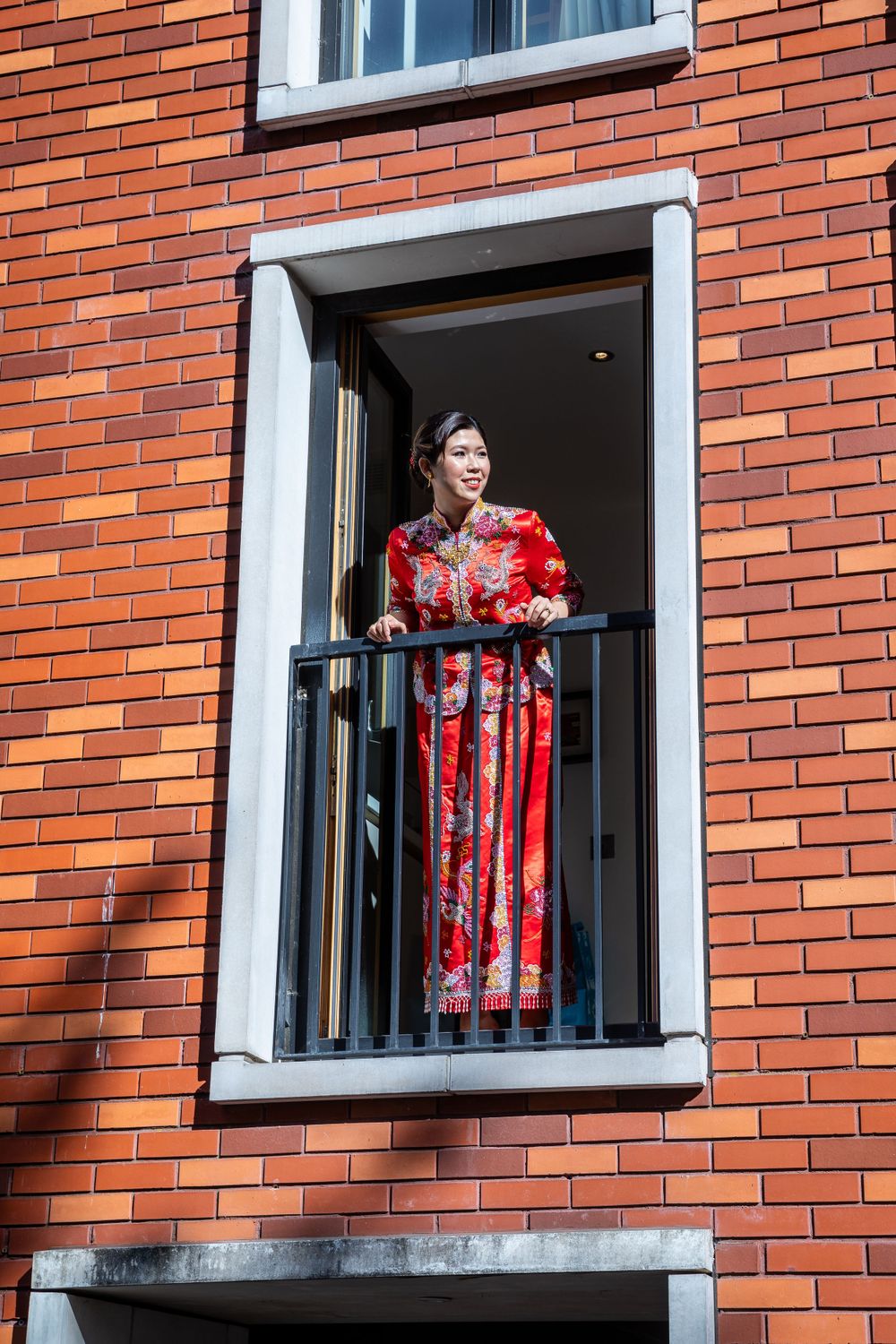 Bride wearing a traditional red Chinese qipao dress standing on a balcony of a red brick building.