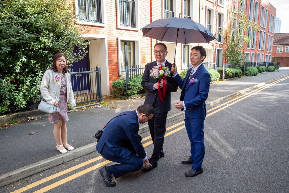 Groom arriving with family members holding umbrellas and flowers outside a brick building for a Chinese wedding.
