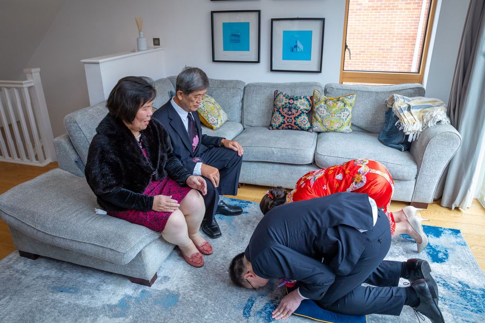 Couple performing traditional Chinese wedding tea ceremony bow before seated elders in a modern living room.