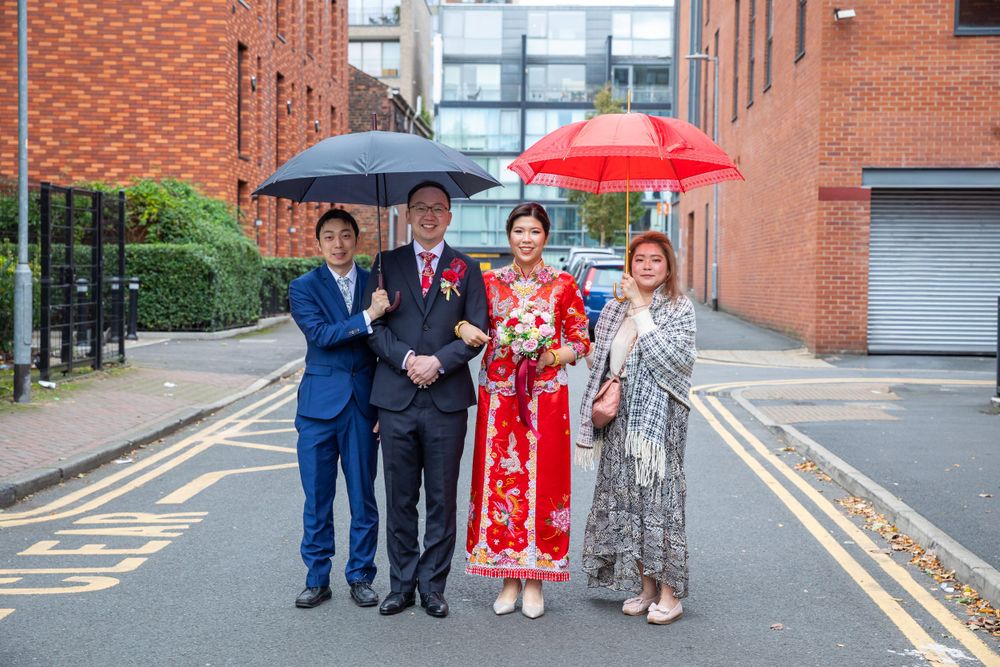 Bride in traditional red Chinese wedding dress posing with family members on a street outside brick buildings.