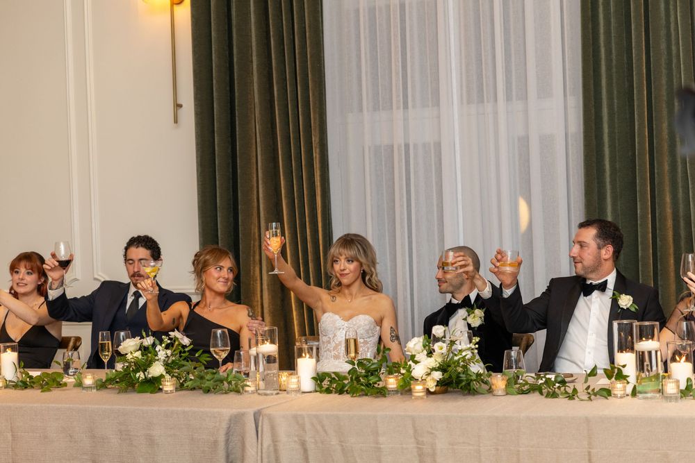 Wedding reception guests seated at long tables under warm lighting in an elegant ballroom setting.