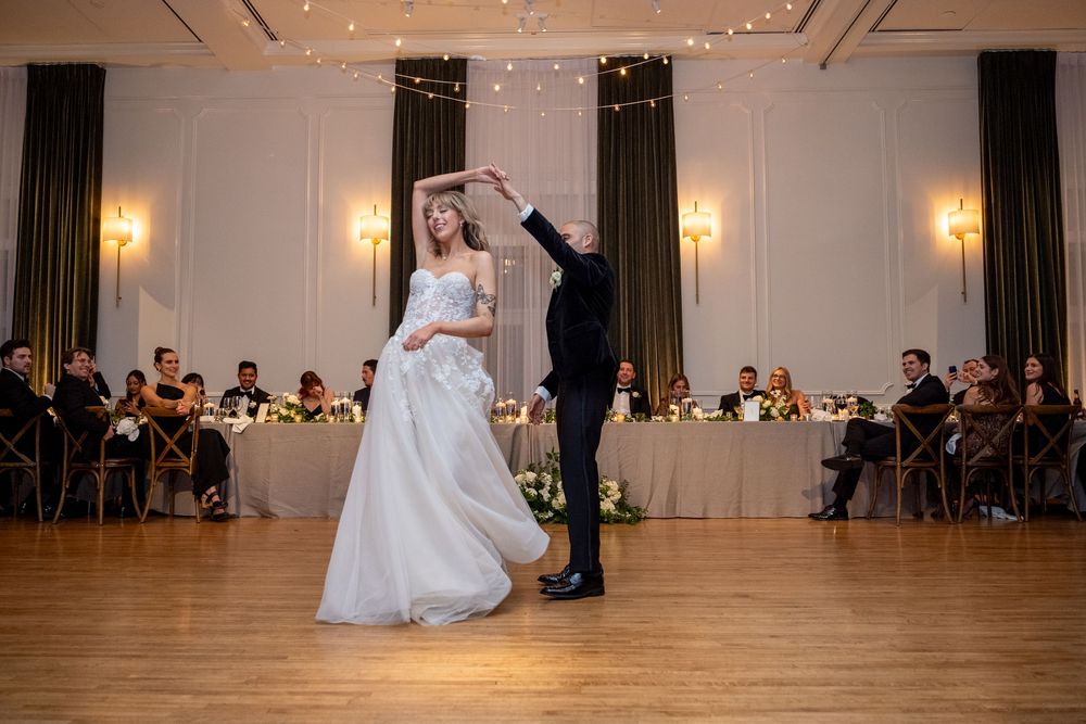 A couple shares their first dance in an elegant ballroom with warm lighting and guests seated at tables in the background.