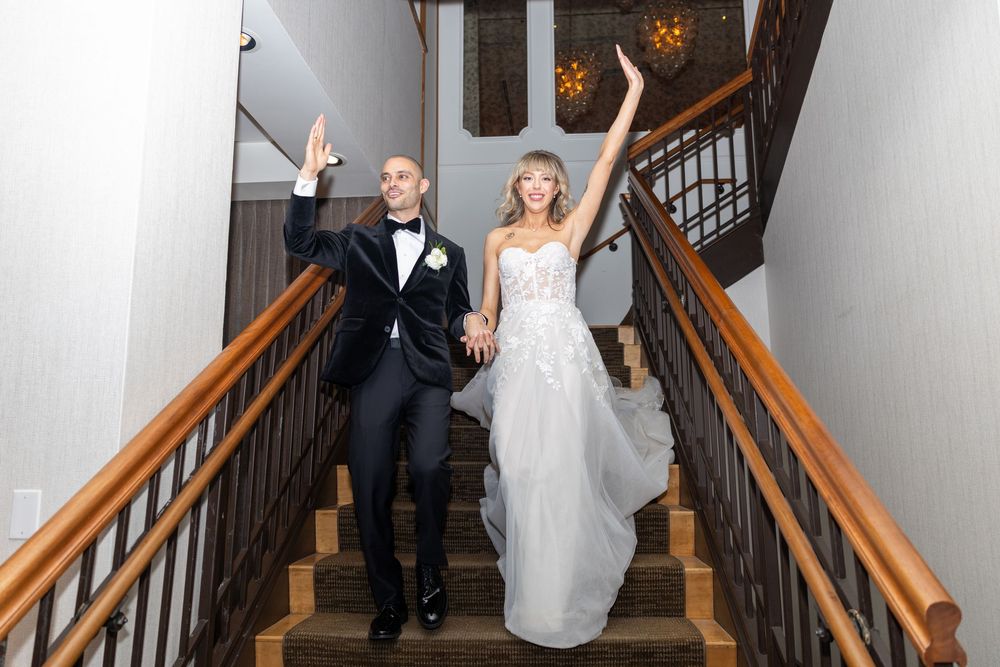 A bride and groom descend an elegant wooden staircase in a rustic venue during their wedding celebration.