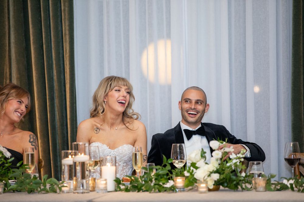 Wedding party seated at a long head table with decorative greenery and candlelight.