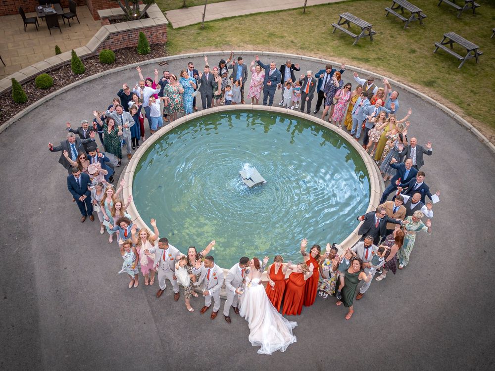 Aerial view of wedding guests forming circle around fountain during outdoor celebration.