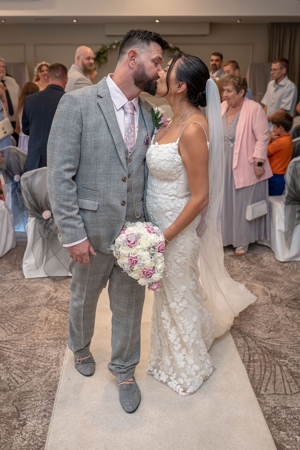 Wedding couple in grey suit and lace dress posing together on patterned carpet.