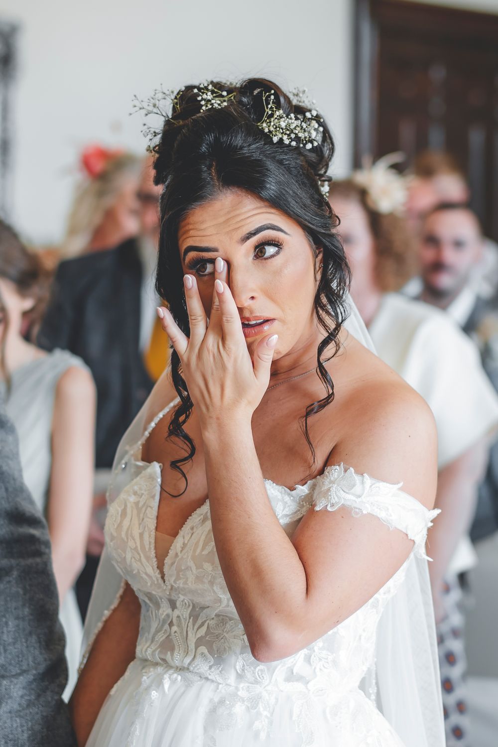 Emotional bride in white lace dress wipes tears during ceremony.