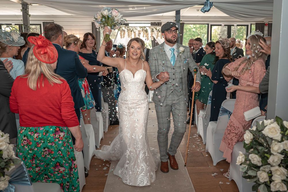Newlyweds walk down aisle while guests celebrate in decorated indoor venue.