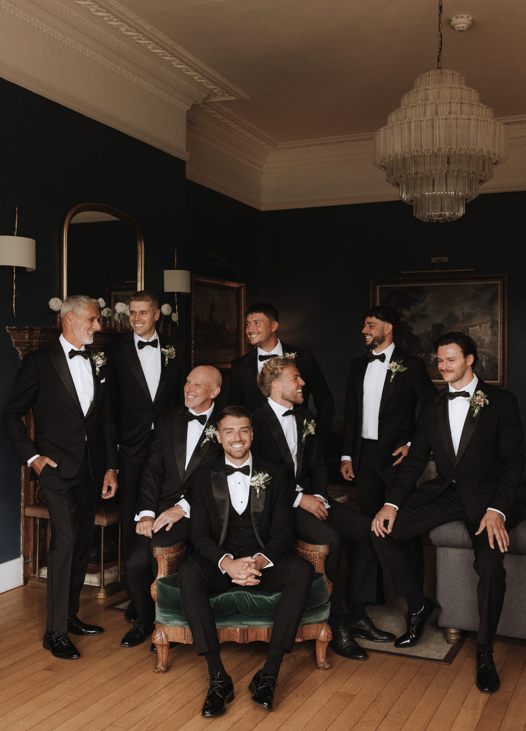 Groomsmen in black tuxedos and bow ties pose together in a formal wedding party lineup against a dark wall at Deer Park Country House.