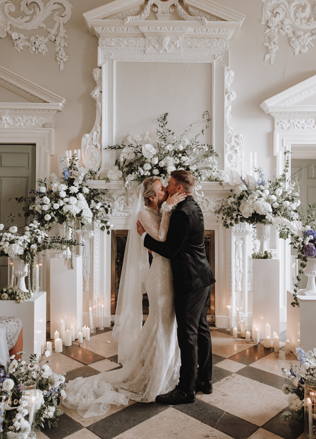 Elegant wedding couple embraces in a grand white hallway adorned with ornate moldings and floral arrangements at Crowcombe Court.