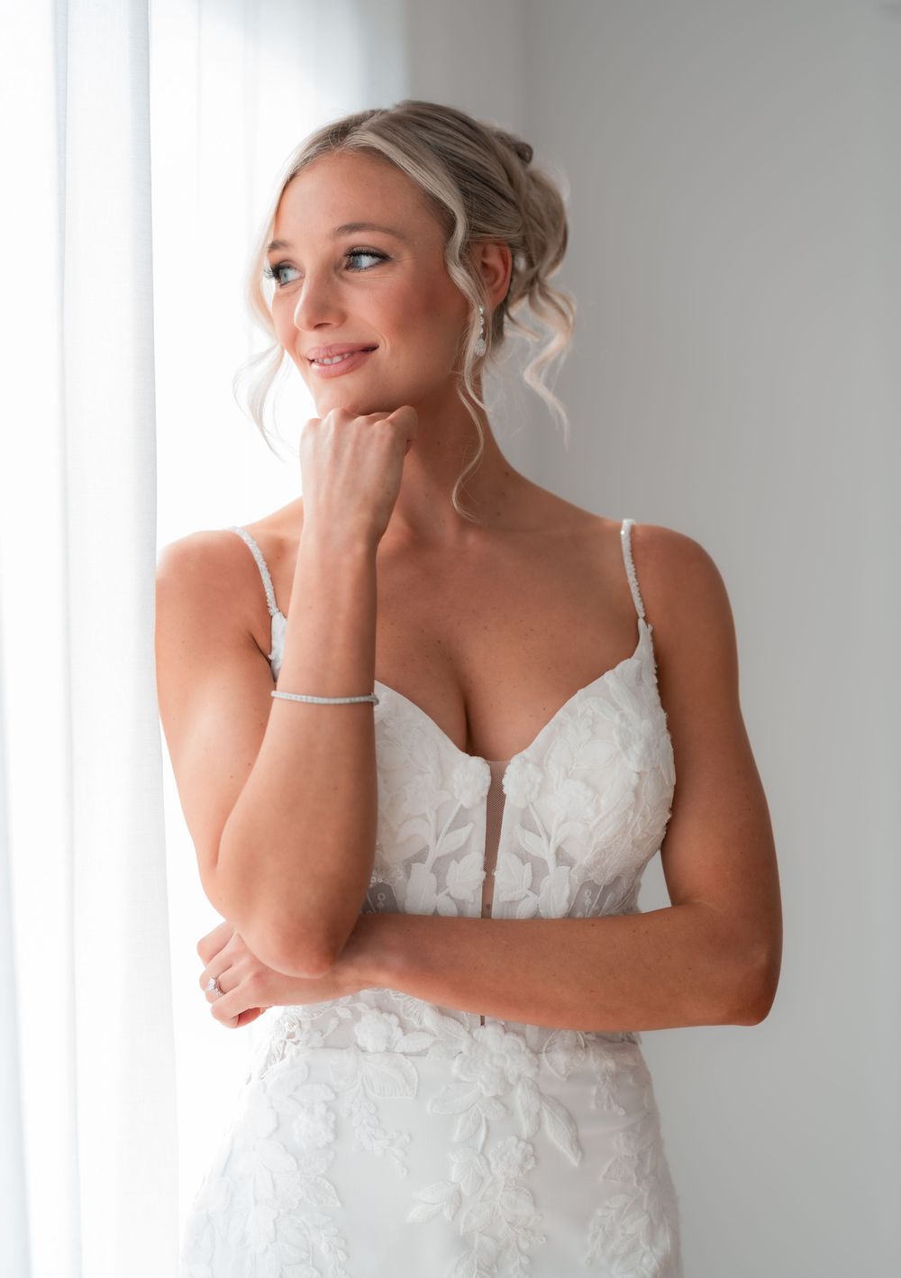 Bride in white lace spaghetti strap wedding gown with updo hairstyle, diamond bracelet, gazing thoughtfully near window.