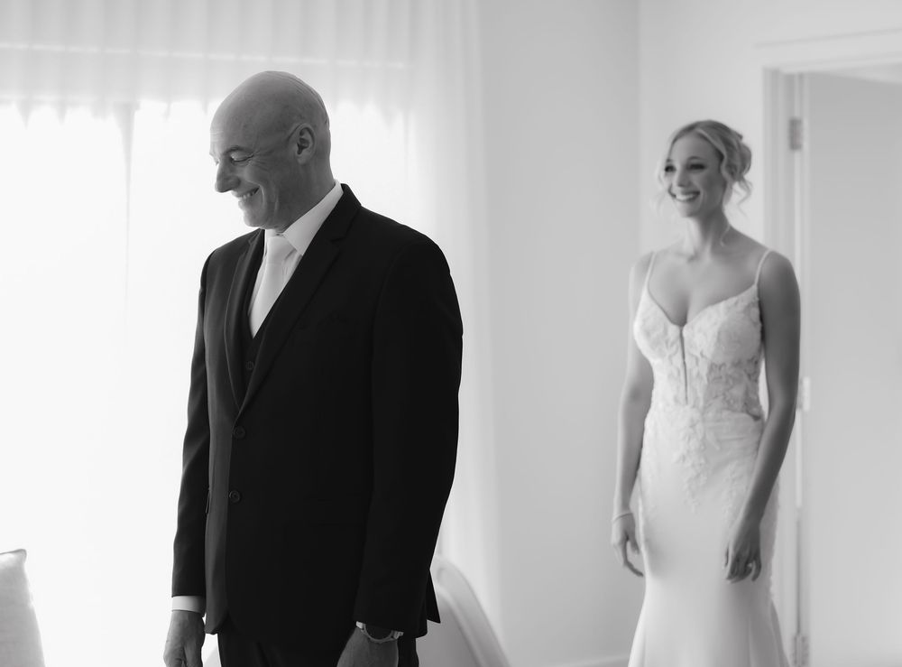 Black and white photo of a bride in a lace gown smiling at a suited man during a wedding first look.